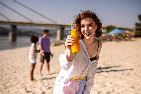Young ginger girl is drinking orange juice at the beach while her friends are hanging out behind her.の写真素材