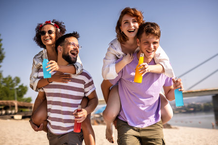 Two male friends are carrying their girl friends on their back on a beach. They are fooling around and drinking cocktails.の写真素材