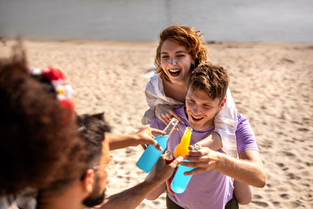 Two male friends are carrying their girl friends on their back on a beach. They are fooling around and drinking cocktails.の写真素材