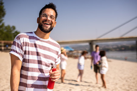 Young handsome man drinking red drink at the beach while his friends are hanging out behind him.の写真素材
