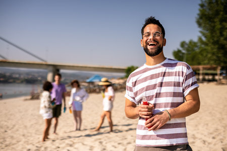 Young handsome man drinking red drink at the beach while his friends are hanging out behind him.の写真素材