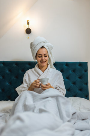 Woman in a hotel room. Wearing bathrobe and a towel on her head. Drinking hot drink after a bath.の写真素材