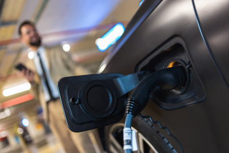 Young businessman using his phone at the charging station while his electric car is charging. Eco electric car conceptの写真素材