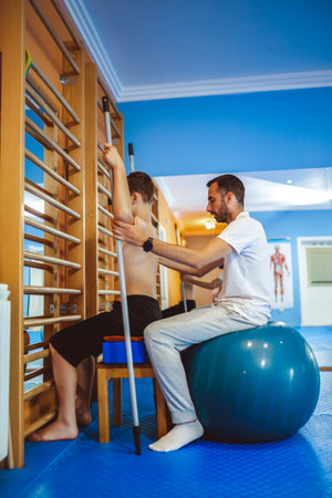 Therapeutic exercise kinesitherapy of little boy. Straightening the pelvis as the boy adheres and sits on exercise ball. A chiropractor is helping him.の写真素材