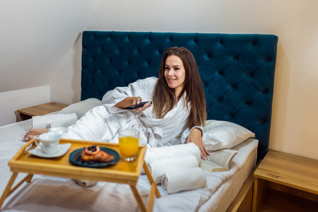 Woman in a bathrobe watching television in her bed on a vacation in a hotel room.の写真素材