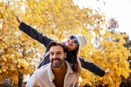 Boyfriend giving girlfriend piggy back ride in a colorful park on a sunny autumn day.