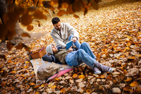 Young cute couple has picnic and reads book together in an colorful autumn park.の写真素材