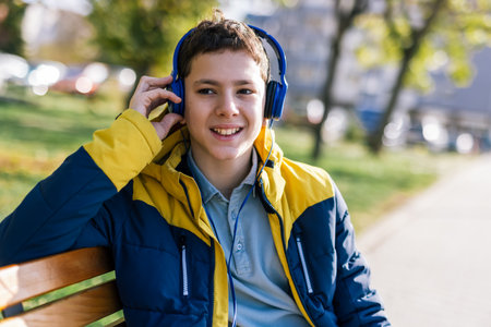 A young boy sitting on a wooden bench in a park, wearing headphones over his ears and dressed in casual clothes.の写真素材