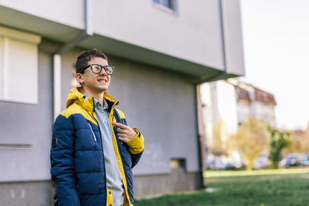 A young boy with glasses walking down a city street wearing a winter jacket and carrying a backpack on his shouldersの写真素材