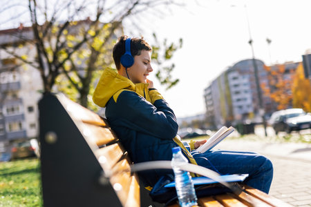A boy sitting on a wooden bench in a park, dressed in casual clothing, wearing headphones over his ears. He is holding an open book, deeply engrossed in readingの写真素材