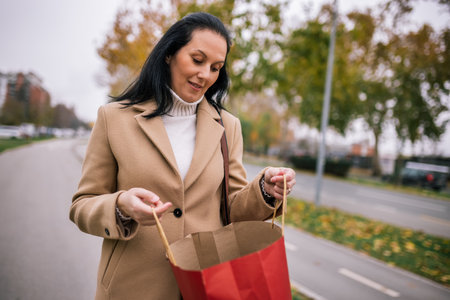 A middle-aged woman walking down the street, wearing a stylish coat and carrying shopping bagsの写真素材