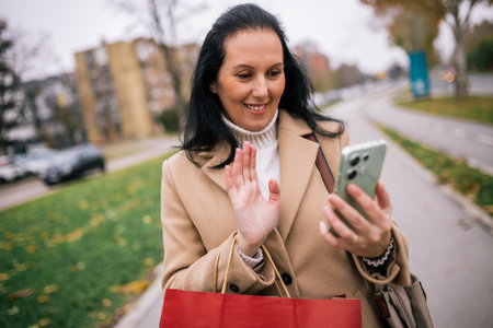 A middle-aged woman walking down the street in a stylish coat, carrying shopping bags while talking on a video callの写真素材