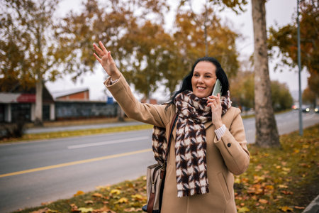 A middle-aged woman in a stylish coat, talking on the phone while signaling for a taxi with her handの写真素材