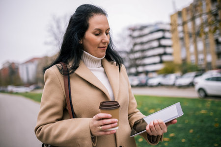 A middle-aged woman in a stylish coat, holding a tablet in one hand and a takeaway coffee cup in the otherの写真素材