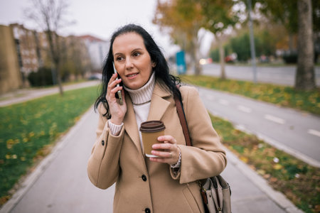 A middle-aged woman in a stylish coat, holding a takeaway coffee cup in one hand while talking on her phone with the otheの写真素材