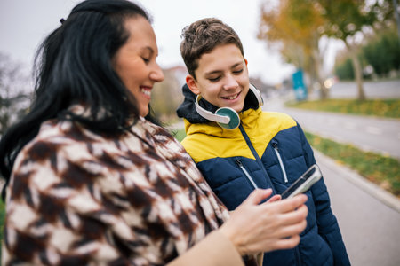 A young boy walking down a city street with his mother, both dressed in warm winter clothing. They are looking at a smartphone together.の写真素材