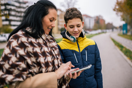 A young boy walking down a city street with his mother, both dressed in warm winter clothing. They are looking at a smartphone together.の写真素材