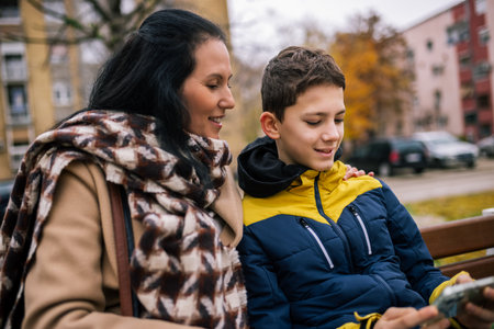 A middle-aged woman in a stylish coat sitting on a bench with her son. Both are looking at a smartphone together, sharing a moment of connection.の写真素材