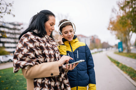 A young boy walking down a city street with his mother, both dressed in warm winter clothing. They are looking at a smartphone together.の写真素材