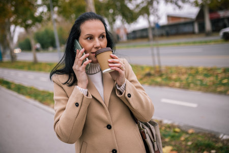 A middle-aged woman in a stylish coat, holding a takeaway coffee cup in one hand while talking on her phone with the otheの写真素材