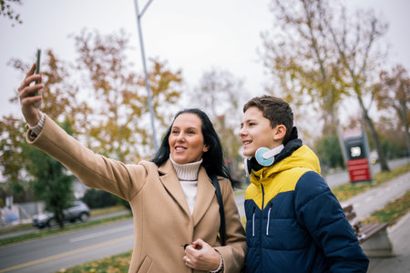 A young boy walking down a city street with his mother, both dressed in warm winter clothing, taking a selfie togetherの写真素材