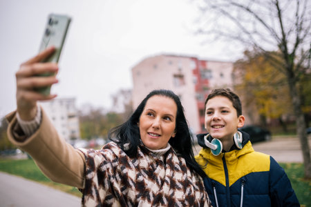 A young boy walking down a city street with his mother, both dressed in warm winter clothing, taking a selfie togetherの写真素材