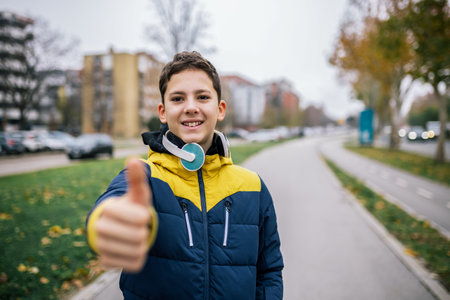 A young boy walking down a city street, wearing headphones and dressed in warm winter clothing. He is giving a thumbs-up with one hand while strolling casually along the sidewalkの写真素材