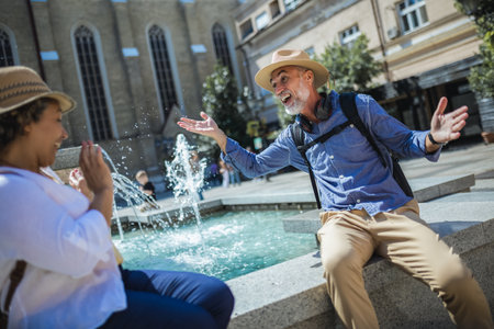 Senior couple of tourists stands happily in front of a stunning city fountain, surrounded by historic architectureの写真素材
