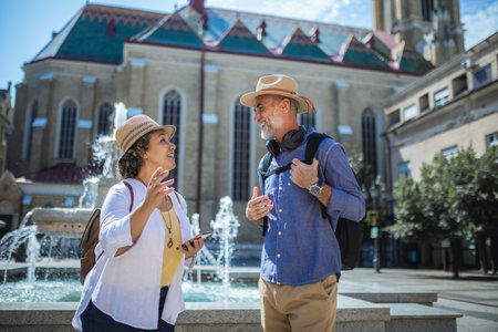 Senior couple of tourists stands happily in front of a stunning city fountain, surrounded by historic architectureの写真素材