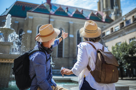 Senior couple of tourists stands happily in front of a stunning city fountain, surrounded by historic architectureの写真素材