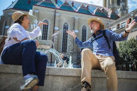 Senior couple of tourists stands happily in front of a stunning city fountain, surrounded by historic architectureの写真素材