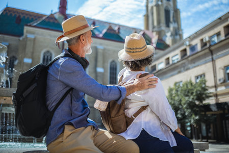 Senior couple of tourists stands happily in front of a stunning city fountain, surrounded by historic architectureの写真素材