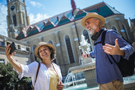 Senior couple of tourists stands happily in front of a stunning city fountain, surrounded by historic architectureの写真素材