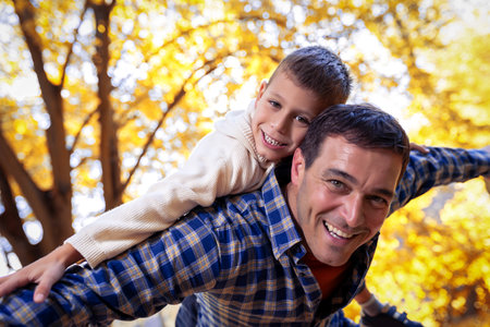 Smiling father gives his young son a piggyback ride on his back in autumn park.の写真素材