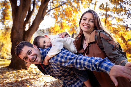Smiling father gives his young son a piggyback ride on his back in autumn park. Mother is smiling. Family in the park.の写真素材