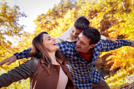 Smiling father gives his young son a piggyback ride on his back in autumn park. Mother is smiling. Family in the park.の写真素材