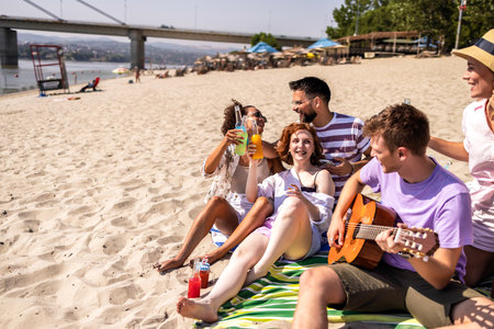 A group of young friends relaxing on the beach during a warm summer day. One guy is playing the guitar while the others enjoy colorful cocktails, laugh, and soak in the sun.の写真素材