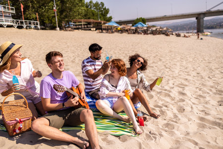 A group of young friends relaxing on the beach during a warm summer day. One guy is playing the guitar while the others enjoy colorful cocktails, laugh, and soak in the sun.の写真素材