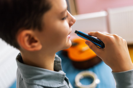 A boy holds a small harmonica to his mouth, playing it while seated on his bed.