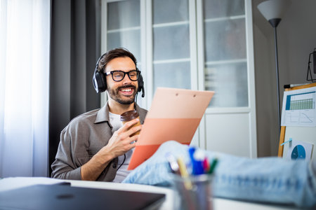 A young man working or studying from home, listening to music with headphones, feet up on the table, and writing in a notebook.の写真素材