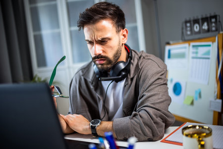 A tired young man working on his laptop at home, with headphones resting around his neck.の写真素材
