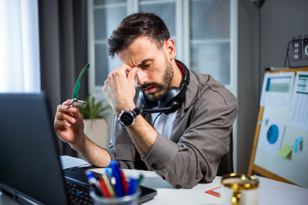 A tired young man working on his laptop at home, with headphones resting around his neck.の写真素材