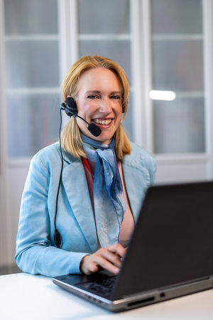 A woman wearing headphones with a microphone, focused on her work while using a laptop. The image...