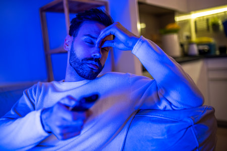A sleepy young man watches TV in a dimly lit room, his eyes heavy with fatigue.の写真素材