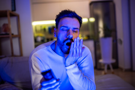 A sleepy young man watches TV in a dimly lit room, his eyes heavy with fatigue.の写真素材