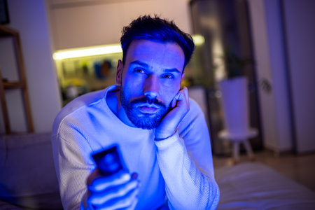 A sleepy young man watches TV in a dimly lit room, his eyes heavy with fatigue.の写真素材