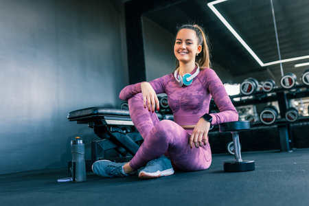Portrait of young fit girl sitting in the gym with headphones and water bottle. She rests from her training.の写真素材