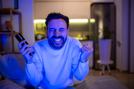 A joyful man watching TV in a dark room, face lit by the soft glow of the screen.の写真素材