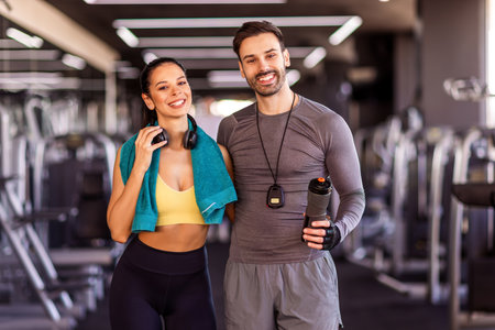 Two gym partners pose to the camera in the gym with towel, headphones and water bottles.の写真素材
