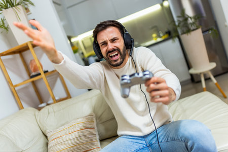 A young man is sitting on a couch in the living room, wearing headphones and holding a game controller as he plays video games.の写真素材